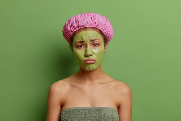 Niezadowolona modelka ze smutkiem patrzy na kamerę nakłada maskę oczyszczającą na wydymane usta usta nosi kapelusz kąpielowy miękki ręcznik wokół nagiego ciała odizolowanego na zielonej ścianie