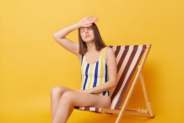 Niezadowolona, zmęczona młoda kobieta, ubrana w strój kąpielowy w paski, siedząca na leżaku odizolowana na żółtym tle, mająca letnie wakacje, kładąc ręce na głowie, ma udar słoneczny