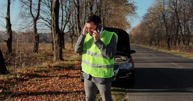 Niezadowolony biznesmen w kamizelce odblaskowej stojący rozmawia przez telefon w pobliżu zepsutego samochodu otworzył maskę pomoc drogową naprawę.
