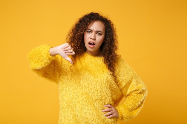 Niezadowolony młody african american dziewczyna w futro sweter pozowanie na białym tle na żółtym pomarańczowym tle, portret studio. Koncepcja życia szczere emocje ludzi. Makieta miejsca na kopię. Pokazuje kciuk w dół.
