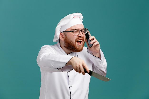 Niezadowolony młody szef kuchni w okularach i czapce rozmawia przez telefon, patrząc na bok wskazujący nożem na bok na białym tle na niebieskim tle