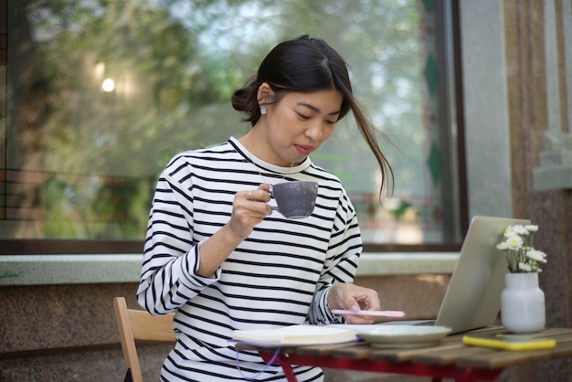 Niezależna dziewczyna pracująca przy komputerze, pisząca w notesie i pijąca kawę w kawiarni na świeżym powietrzu