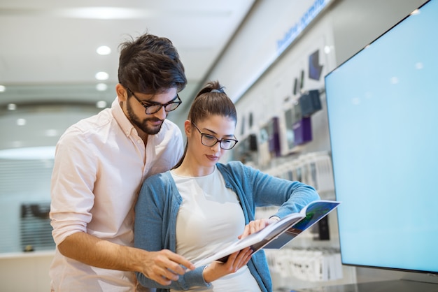 Niezdecydowana młoda atrakcyjna para robi zakupy w sklepie elektronicznym, przeglądając katalog produktów.