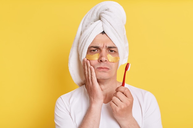 Niezdrowy mężczyzna owinięty ręcznikiem na głowie wykonujący poranne zabiegi kosmetologiczne odczuwa ból po umyciu zębów pozowanie z łatami pod oczami na białym tle na żółtym tle