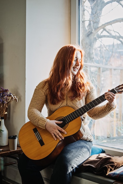 Niezmieniony Szczery Portret Młodej Rudowłosej Kobiety W Swetrze Grającej Na Gitarze Akustycznej, Siedzącej Obok