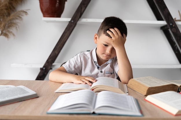 Niezmotywowany uczeń z bólem głowy siedzący w domu przy biurku odrabiający pracę domową przygotowujący się do lekcji Zmęczony mały uczeń szkoły czytający książki uczący się online koncepcja wiedzy na odległość