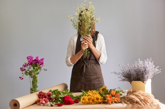 Nieznana anonimowa kobieta kwiaciarnia tworząca piękny bukiet w miejscu pracy w kwiaciarni stojącej na szarym tle, zakrywającej twarz roślinami