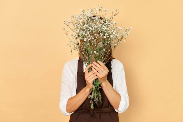 Nieznana nieznana kobieta kwiaciarka w brązowym fartuchu ukrywająca twarz za bukietem kwiatów stojąca odizolowana na beżowym tle