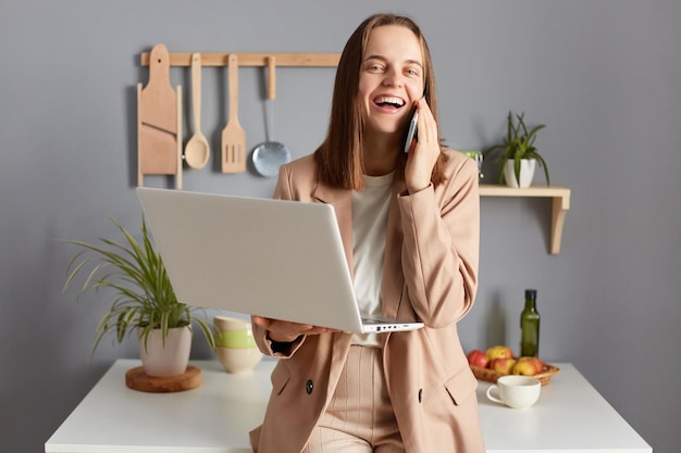 Niezwykle szczęśliwa kobieta o brązowych włosach, ubrana w formalny garnitur, pozuje w kuchni, pracując od szlifowania na notebooku, mając przerwę rozmawiającą na smartfonie