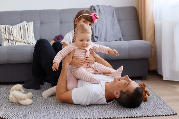 Niezwykle szczęśliwy młody dorosły mężczyzna leżący na podłodze w pobliżu sofy i bawiący się ze swoimi małymi dziećmi, ubrany w białą koszulkę i spodnie, dzieci siedzące na ojcu, rodzina ciesząca się wspólnym spędzaniem czasu.