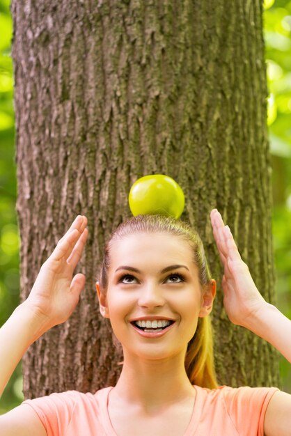 Niosąc jabłko na głowie. Figlarna młoda kobieta niosąca jabłko na głowie i patrząca w górę z uśmiechem, opierając się o drzewo w parku