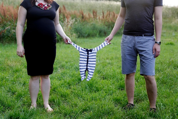 Zdjęcie niska część pary trzymająca ubrania dla niemowląt stojące na trawiastym polu