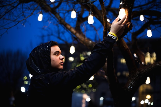 Niski kąt widoku człowieka trzymającego oświetloną żarówkę wiszącą na drzewie w nocy