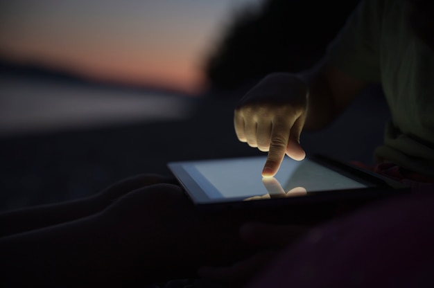Niski kąt widzenia dziecka za pomocą cyfrowego tabletu, siedzącego na plaży wieczorem.