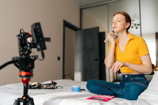 Niski kąt widzenia rudowłosej blogerki piękności, która robi samouczek wideo przy użyciu profesjonalnej kamery stojącej na statywie, mówiącej, jak krok po kroku nakładać makijaż