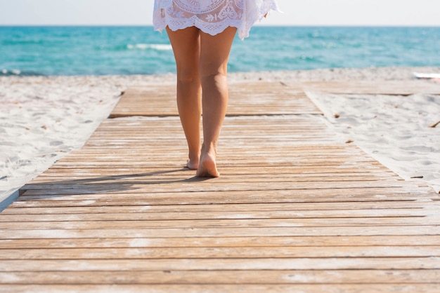 Niski przekrój kobiety chodzącej po drewnianej desce wśród piasku prowadzącej w kierunku wody morskiej. Kobieta idąca w kierunku morza przez drewnianą ścieżkę wzdłuż piaszczystej plaży