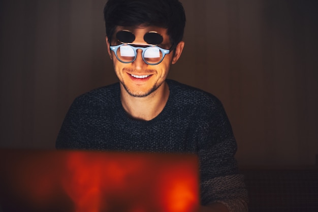 Noc młody szczęśliwy człowiek ubrany w okrągłe okulary, patrząc w laptopa w ciemnym pokoju z girlandami w domu.