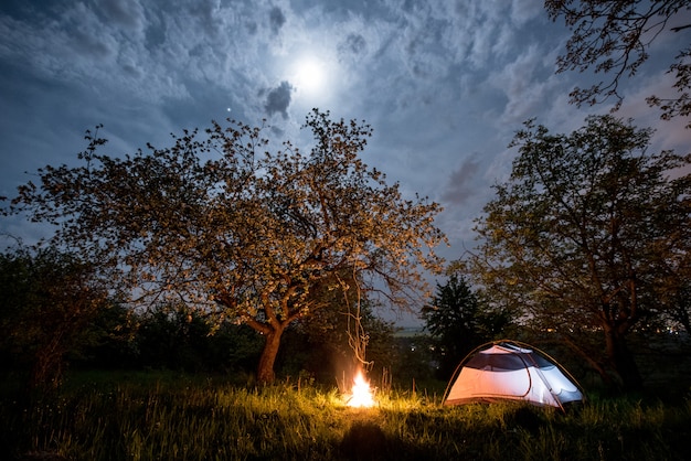 Nocne Biwakowanie. Podświetlany Namiot Turystyczny Przy Ognisku Pod Drzewami I Nocnym Niebem Z Księżycem