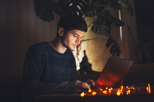 Nocny młody facet wpisując na laptopie w ciemnym pokoju z girlandami na biurku w domu.