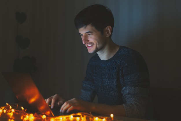 Nocny młody wesoły facet pracuje na laptopie w ciemnym pokoju z girlandami na stole w domu.