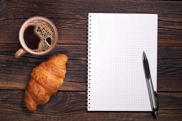 Notatnik długopis i filiżanka kawy z rogalikiem na drewnianym tle Miejsce na tekst