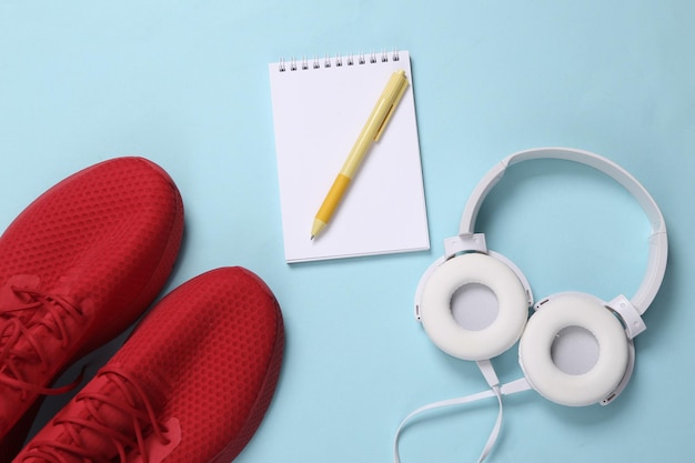 Notebook ze słuchawkami i czerwone buty sportowe na niebieskim tle Plan treningowy Koncepcja sportu Uruchamianie akcesoriów do aktywnego stylu życia Widok z góry
