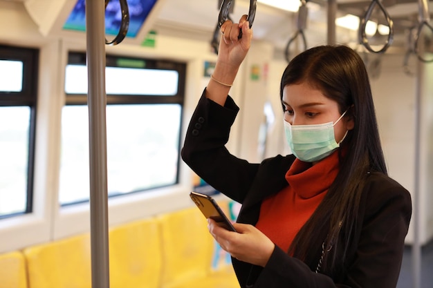 Nowa normalna epidemia Covid-19, młoda biznesowa azjatycka kobieta nosząca maskę ochronną, aby zapobiec zakażeniu wirusem Covid-19 lub koronawirusem od osób w pociągu, korzystając z komunikacji smartfona w pociągu