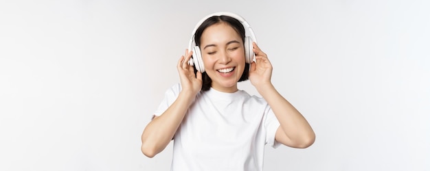 Nowoczesna azjatycka dziewczyna tańczy słuchając muzyki ze słuchawkami, uśmiechając się, szczęśliwa stojąc w koszulce na białym tle