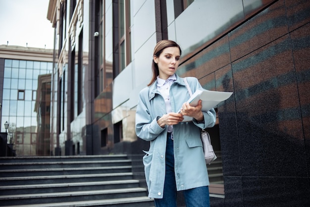 Nowoczesna biznesowa kobieta uważnie przygląda się folderowi z dokumentami na tle budynku biznesowego ze schodami Styl życia