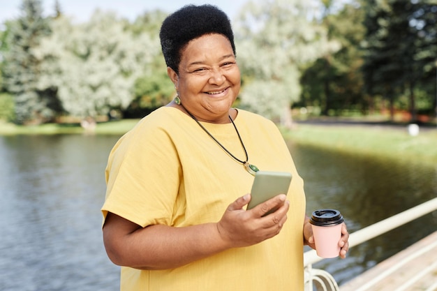 Nowoczesna czarna starsza kobieta używa smartfona na świeżym powietrzu w parku i uśmiecha się