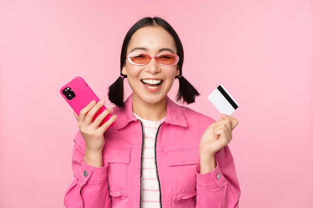Nowoczesna piękna azjatycka dziewczyna śmiejąca się i uśmiechająca się z karty kredytowej telefonu komórkowego zakupy online płacąc smartfonem stojącym na różowym tle
