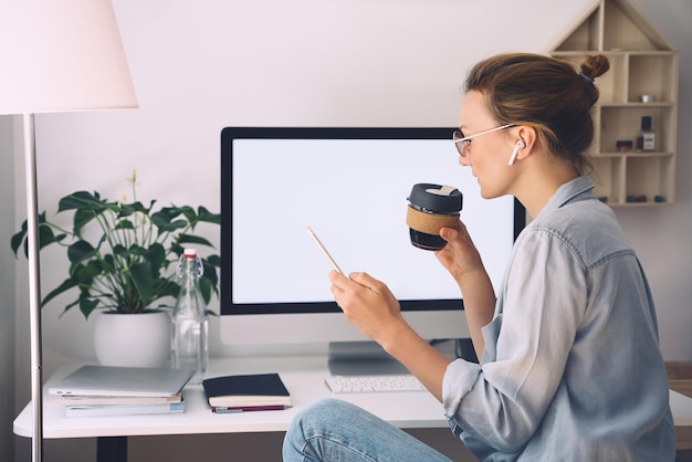 Nowoczesna uśmiechnięta piękna kobieta pracuje i pije kawę z kubka wielokrotnego użytku w domowym biurze Minimalistyczne miejsce pracy w stylu slow life kreatywnej kobiety w okularach i słuchawkach przy użyciu technologii komputerowej