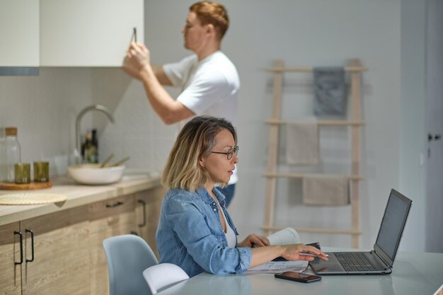 Nowoczesna wielonarodowa para w domu w kuchni kobieta pracująca lub studiująca korzysta z laptopa, mąż w tle gotuje lub sprząta zdrowe relacje bez stereotypów dotyczących płci