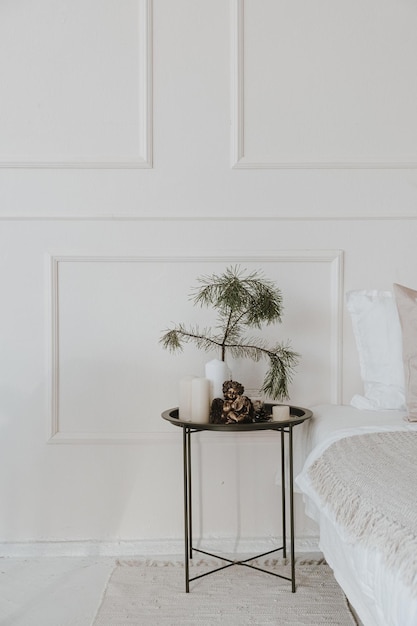 Nowoczesne jasne wnętrze sypialni ze stolikiem nocnym igły jodły gałęzie świec łóżko neutralna pościel poduszki Estetyczny salon z dekoracjami świątecznymi