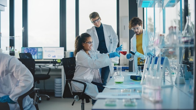 Zdjęcie nowoczesne laboratorium badań medycznych młoda, piękna, czarna studentka nauki mieszanie ciekłej chemii w dwóch pucharach rozmawianie różnorodny zespół specjalistów wieloetnicznych pracuje w zaawansowanym laboratorium