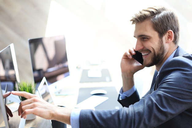 Nowoczesny biznesmen analizowania danych za pomocą komputera i rozmawia przez telefon siedząc w biurze.