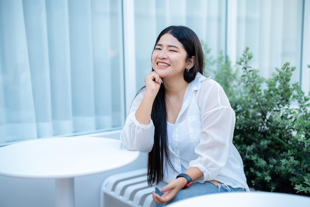 Nowoczesny portret pięknej szczęśliwej azjatyckiej kobiety podróżnika sobie smartwatch na sobie piękną białą sukienkę i nosić dżinsy styl życia, ciesząc się siedzącą kawiarnią restauracja tło