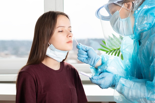 Nowoczesny pracownik laboratorium noszący środki ochrony osobistej testuje młodą kobietę na obecność koronawirusa za pomocą wymazu z nosa