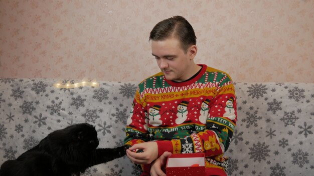 Noworoczny młodzieniec otwiera i pakuje prezent, szampana, prezenty noworoczne, noworoczne dekoracje i dobry nastrój oraz czarny kot
