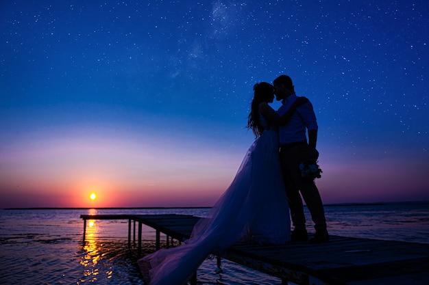 Nowożeńcy na oceanie o zachodzie słońca. tylko zakochana para, która świętowała ślub w sukniach ślubnych, stoi na drewnianym moście na morzu na tle pięknego zachodu słońca.