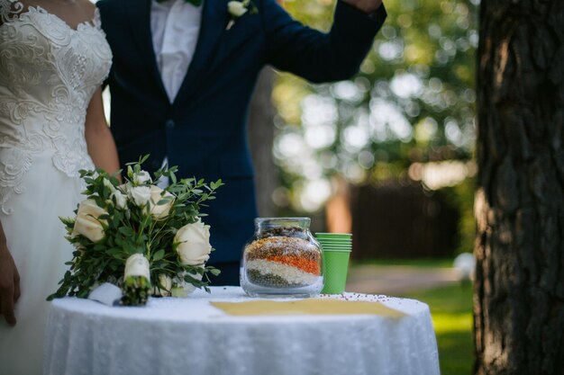 Nowożeńcy przytulający się przy uroczym stoliku ze słodyczami, za nimi zieleń. nieostrość