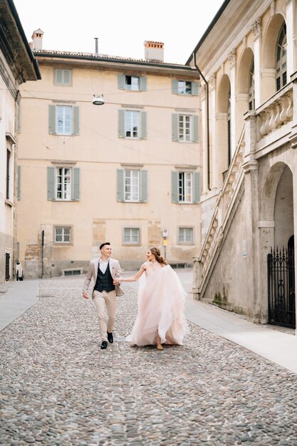 Nowożeńcy trzymający się za ręce chodzący po bruku w pobliżu starego budynku w bergamo we włoszech