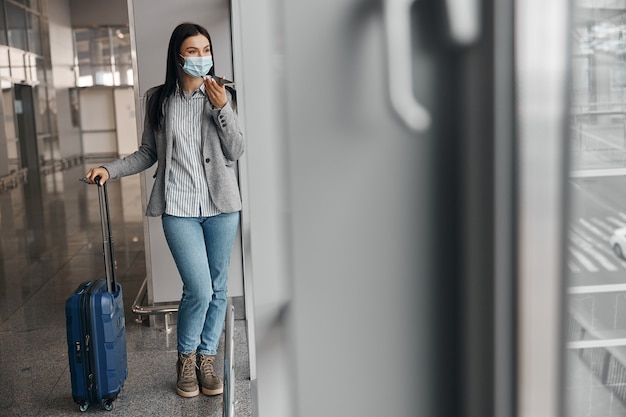 Nowy porządek na lotniskach. Kobieta w masce w terminalu z bagażem w pobliżu panoramicznych okien sama
