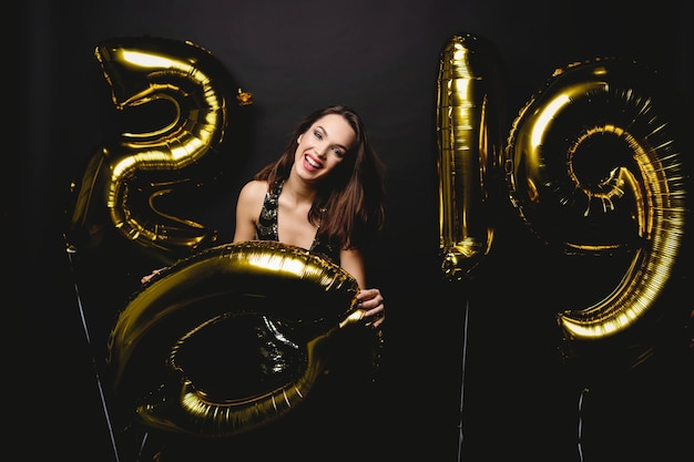 Nowy Rok. Kobieta Z Balonami Świętuje Przy Przyjęciem. Portret pięknej uśmiechniętej dziewczyny w błyszczącej sukience rzucanie konfetti, zabawy z balonami złota 2019 na tle. Wysoka rozdzielczość.