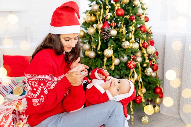Nowy Rok lub Boże Narodzenie, młoda mama z dzieckiem na łóżku w domu przy choince w stroju Świętego Mikołaja przytulająca się i czekająca na święta uśmiechnięta