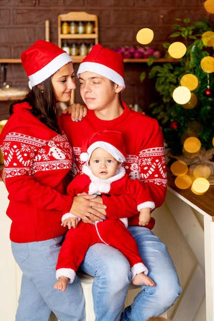 Nowy Rok lub Boże Narodzenie, szczęśliwa młoda rodzinna mama, tata i dziecko w ciemnej kuchni domu z choinką w czerwonych swetrach i czapkach Świętego Mikołaja uśmiechający się, przytulający się, gratulujący święta