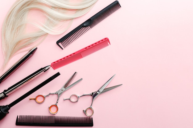 Nożyczki, grzebienie, prostownica do włosów i inne akcesoria fryzjerskie oraz kosmyk blond włosów na różowym tle. Usługi fryzjerskie.
