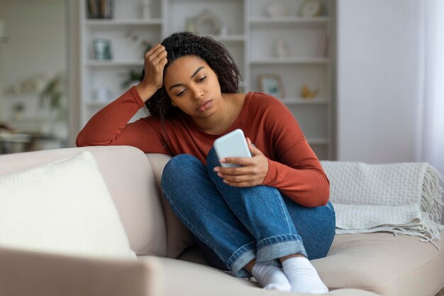 Nudzona młoda czarna kobieta siedząca na kanapie i patrząca na ekran smartfona