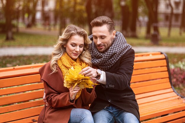 Obejmująca szczęśliwa para siedząca delikatnie romantycznie przytulona na ławce w parku w płaszczach i szalikach