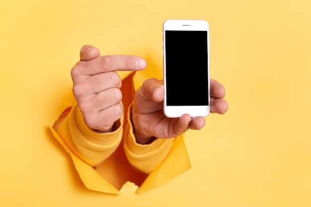 Obraz dłoni osoby przebijającej się przez żółty papier i trzymającej smartfona wskazującego na pusty ekran z miejscem na kopię na tekst reklamowy lub promocyjny